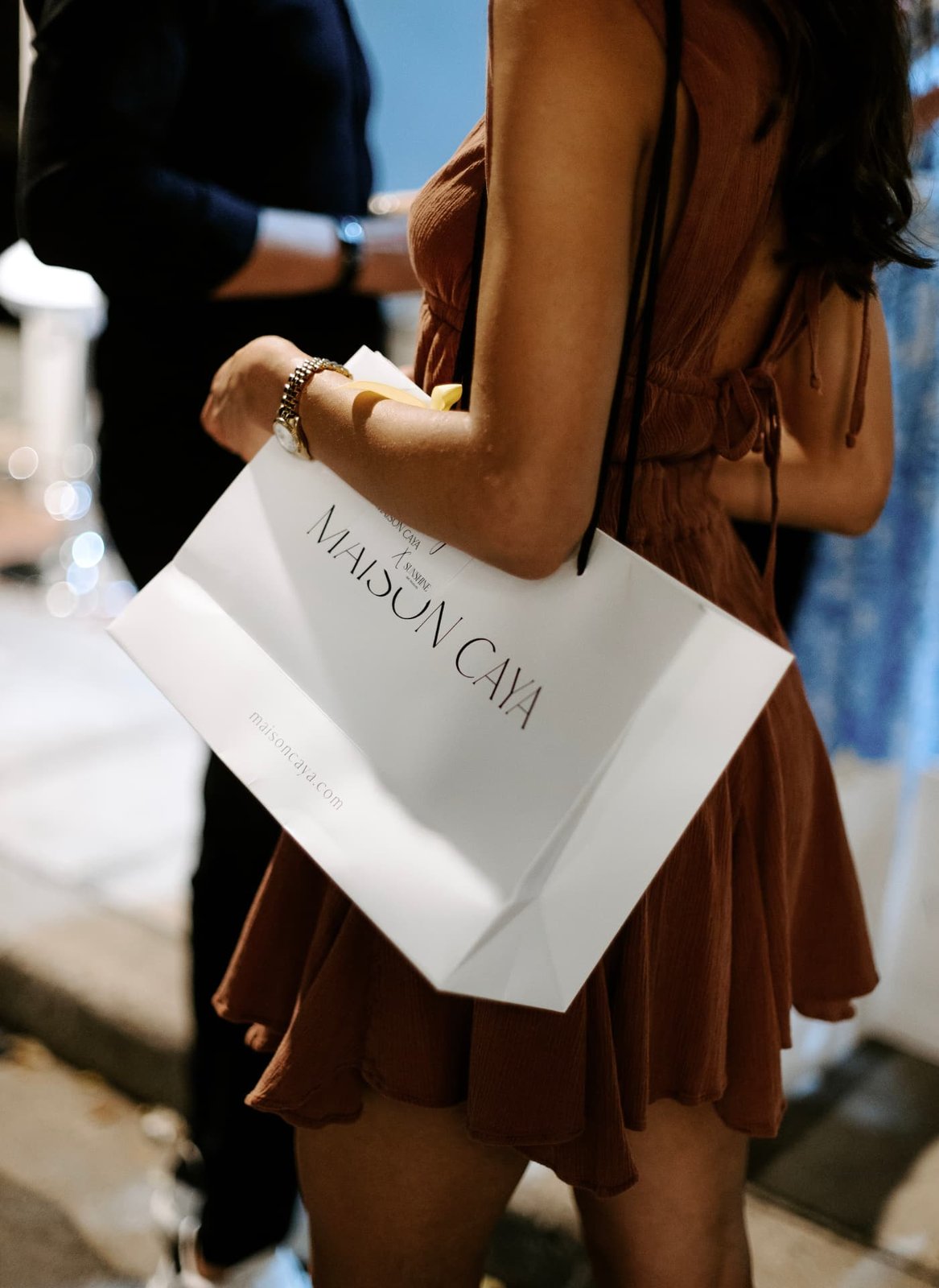 Guest in a bronze dress carrying a white Maison Caya shopping bag